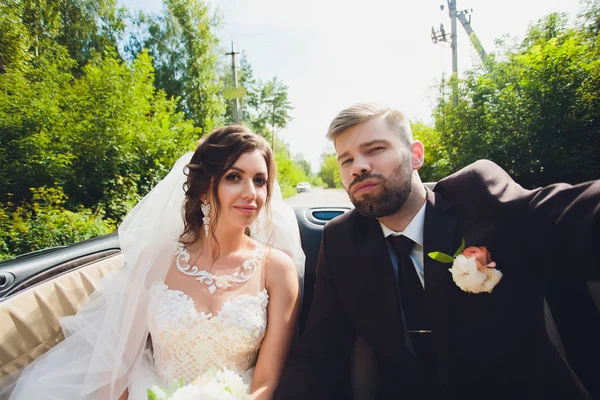 Närbild porträtt ung vacker snygg par brud i en vit klänning med en bukett blommor i håret och och brudgummen i bilen på vägen. — Stockfoto