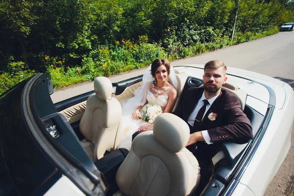 Портрет крупным планом молодая красивая стильная пара невеста в белом платье с букетом цветов в волосах и жених в машине по дороге . — стоковое фото
