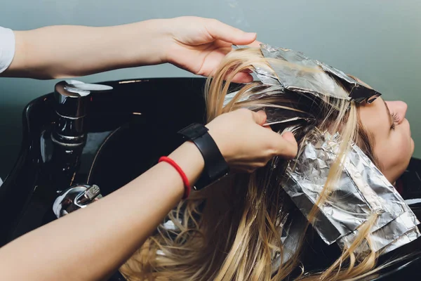 Închiderea mâinilor coafor care separă firele de păr ale unei femei tinere frumoase cu folie de aluminiu înainte de a schimba culoarea părului . — Fotografie, imagine de stoc