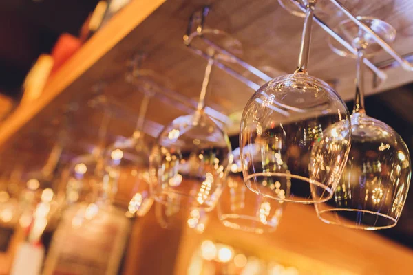 Closeup many upside down empty clear transparent crystal upturned wine glasses hanging in straight rows on brown wooden shelf, rack, showcase, stainless steel railing in bar, cabinet.