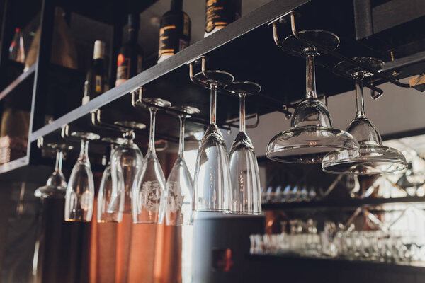 Closeup many upside down empty clear transparent crystal upturned wine glasses hanging in straight rows on brown wooden shelf, rack, showcase, stainless steel railing in bar, cabinet.