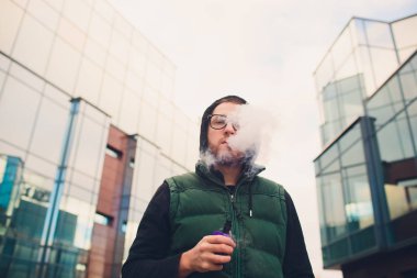 Geniş sakallı, gözlüklü bir gencin portresi şehrin arka planında elektronik bir sigara içiyor..