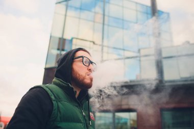 Geniş sakallı, gözlüklü bir gencin portresi şehrin arka planında elektronik bir sigara içiyor..