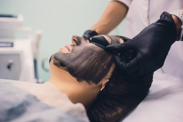 Pembe eldivenli kozmetolog, kozmetoloji odasında genç bir kızın yüzünü soymak için karbon maske takıyor. Kozmetik hizmetleri ve kişisel bakım kavramı. — Stok fotoğraf