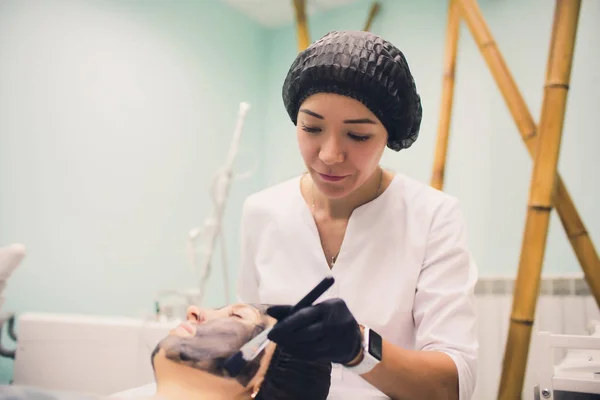 Kosmetolog w różowych rękawiczkach ze szczotką nakłada maskę węglową do obierania twarzy młodej dziewczyny w gabinecie kosmetycznym. Koncepcja usług kosmetologicznych i samoopieki. — Zdjęcie stockowe