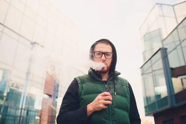 Retrato de un joven con barba grande en gafas vapeando un cigarrillo electrónico frente al fondo urbano . — Foto de Stock
