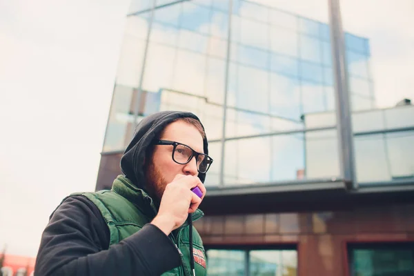 Retrato de un joven con barba grande en gafas vapeando un cigarrillo electrónico frente al fondo urbano . — Foto de Stock