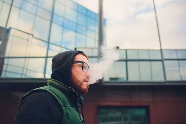 戴着大胡子戴眼镜的年轻人在对面城市背景的电子香烟上抽烟的画像. — 图库照片
