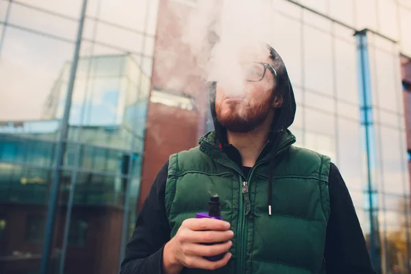 Porträtt av ung kille med stort skägg i glas ånga en elektronisk cigarett motsatt urban bakgrund. — Stockfoto