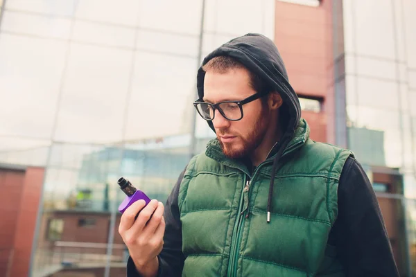 Retrato de un joven con barba grande en gafas vapeando un cigarrillo electrónico frente al fondo urbano . — Foto de Stock