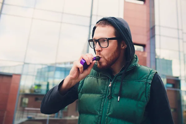 Retrato de un joven con barba grande en gafas vapeando un cigarrillo electrónico frente al fondo urbano . — Foto de Stock