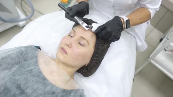 Ο κοσμητολόγος με τα ροζ γάντια με το πινέλο εφαρμόζει μια μάσκα άνθρακα για ξεφλούδισμα στο πρόσωπο ενός νεαρού κοριτσιού σε ένα δωμάτιο κοσμετολογίας. Η έννοια των υπηρεσιών κοσμετολογίας και αυτοφροντίδας. — Αρχείο Βίντεο