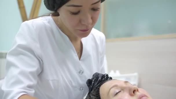 Косметолог в розовых перчатках с кистью наносит углеродную маску для пилинга на лицо молодой девушки в косметологической комнате. Концепция косметологических услуг и самообслуживания . — стоковое видео