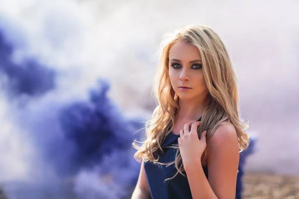 Young beautiful blond woman in blue mini dress and floral wreath standing and trying water on summer day with coutryside landscape at background. Woman natural beauty and summer nature concept. smoke — Stock Photo, Image