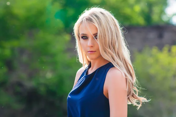 Junge schöne blonde Frau in blauem Minikleid und Blumenkranz steht und probiert Wasser an einem Sommertag mit Landschaft im Hintergrund. Natürliche Schönheit der Frau und sommerliches Naturkonzept. — Stockfoto