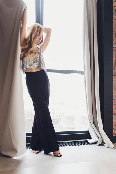 Bela mulher loira em roupas pretas com saltos fica à janela. vista traseira . — Fotografia de Stock