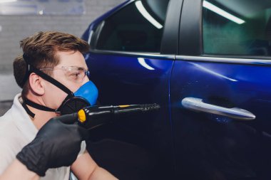 Koruyucu bir giysinin içindeki dezenfektan ve maske dışarıda arabanın dezenfektanlarını spreyliyor. Koronavirüs salgını.