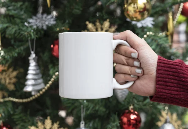 Women hand holding white ceramic coffee cup on christmas tree background. mockup for creative advertising text message or promotional content. — Stock Photo, Image