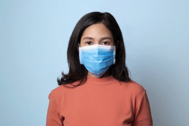 Kadın koruyucu maske takıyor ve mavi arka planda kameraya bakıyor..