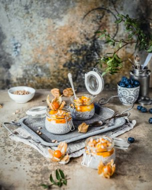 Chia tohumlu puding ve çam fıstıklı mango dilimleri ve pejmürde bej arka plandaki kavanozlarda physalis, sağlıklı kahvaltı konsepti.