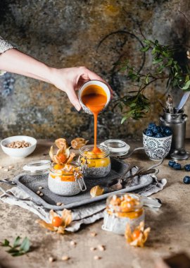 Kadın eli kavanoza mango dolduruyor puding ve meyve dilimli chia tohumu ve pejmürde bej arka planda çam fıstığı, sağlıklı kahvaltı kavramı.