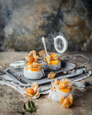 Chia tohumlu puding ve çam fıstıklı mango dilimleri ve pejmürde bej arka plandaki kavanozlarda physalis, sağlıklı kahvaltı konsepti.