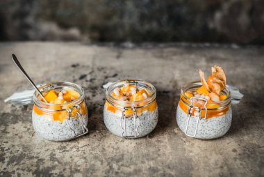 Chia tohumlu puding ve çam fıstıklı mango dilimleri ve pejmürde bej arka plandaki kavanozlarda physalis, sağlıklı kahvaltı konsepti.