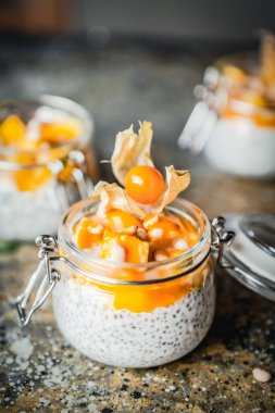 Chia tohumlu puding ve çam fıstıklı mango dilimleri ve pejmürde bej arka plandaki kavanozlarda physalis, sağlıklı kahvaltı konsepti.