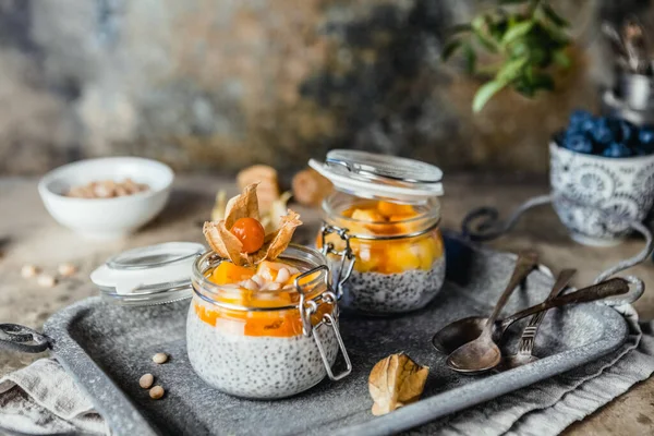 Chia tohumlu puding ve çam fıstıklı mango dilimleri ve pejmürde bej arka plandaki kavanozlarda physalis, sağlıklı kahvaltı konsepti.