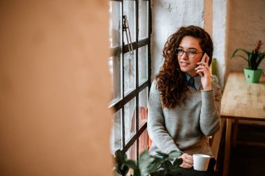 Kahve içme ve cep telefonuyla konuşurken pencere yanında oturan neşeli genç kadın görüntüsünü. 