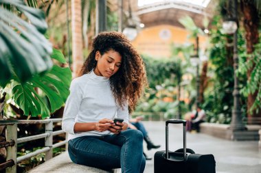 Beaituful kadın turist smartphone kullanarak kıvırcık saçlı. Açık havada bavulla oturan.