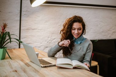 Modern daire ve kitap okumak içinde laptop ile masa başında oturan kıvırcık saçlı kadın portresi.