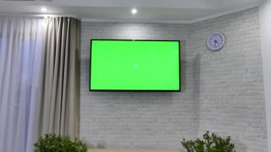 Oturma odasındaki duvarda yeşil ekran bir televizyon asılı. Tuğla bir duvarın arkasında.