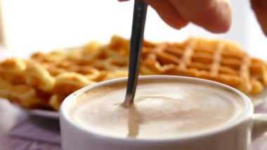 Waffle 'lı sabah kahvesi. Köpüklü bir içeceği kaşıkla bardağa koy. Sütlü ve köpüklü kahve..