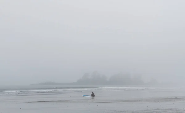 Surfista de fato de mergulho sentado na praia em nevoeiro olhando para o oceano — Fotografia de Stock