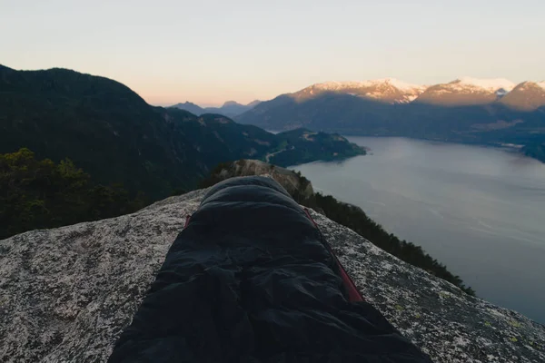 Homem em saco de dormir no topo das montanhas acima do lago, enquanto o sol — Fotografia de Stock