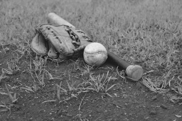 Old baseball bat with ball and glove in black and white lay on field.