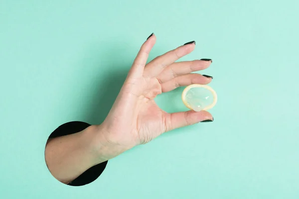 Female hands hold condom through a hole. Safe anonymous sex concept. Isolated on neon mint background.