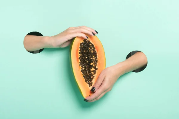 Female hands hold fresh papaya through a hole on neon mint background. Minimalistic creative isolated concept.