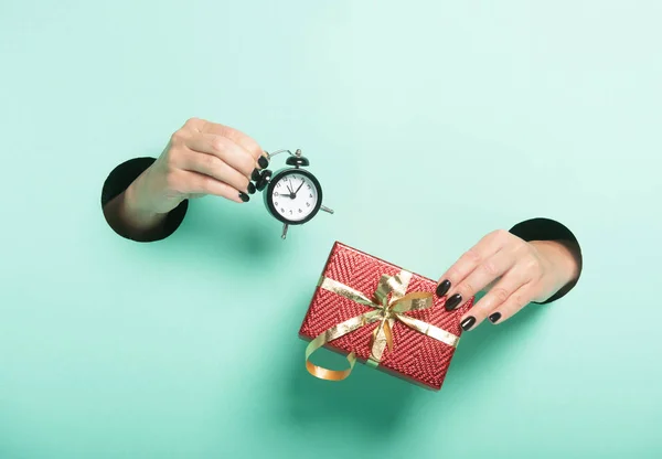 Female hands hold red gift and alarm clock through a hole on neon mint background. Minimalistic creative isolated sale concept. — Stock Photo, Image