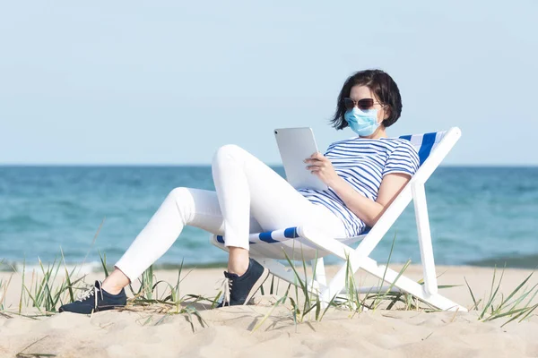 Jonge vrouw in een masker met laptop op lege kust, beschermende uitrusting tijdens coronavirus covid19. Sociaal afstandconcept. Stockfoto