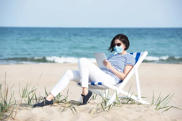 Young woman in a mask sitting with laptop on empty coast, protective equipment during coronavirus covid19. Social distance concept. — Stock Photo, Image