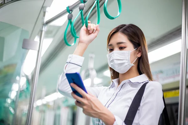 亚洲女商人在乘地铁或火车上下班时 使用带有外科口罩的智能手机进行面部保护 并保持社交距离 — 图库照片