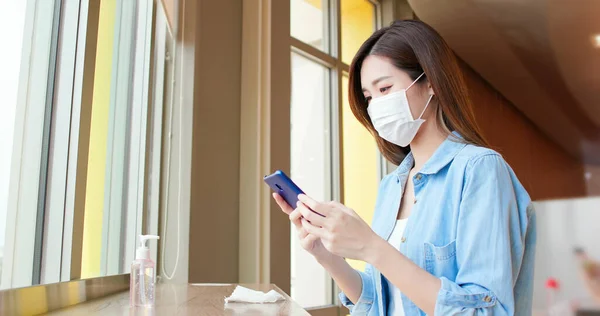 フェイスマスクを身に着けている若いアジアの女性は 彼女が彼女の手をきれいにした後 スマートフォンを使用し 乾燥させるために消毒剤アルコールジェルでテーブルを拭く ウイルスを殺すための概念は Covid 19の普及を防ぐ — ストック写真