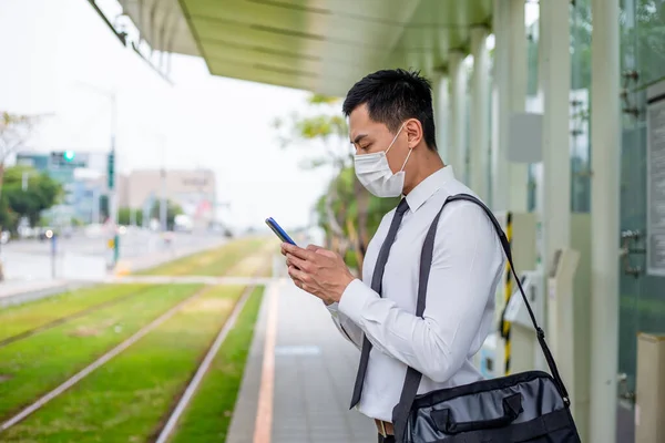 アジアのビジネスマンは 地下鉄や電車を待っている間 外科用マスク顔保護付きスマートフォンを使用しています — ストック写真