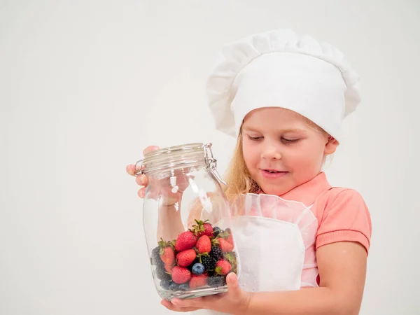 A girl in an apron and a chef\'s hat with a jar in her hands. There are many different berries in the jar. Positive emotions.