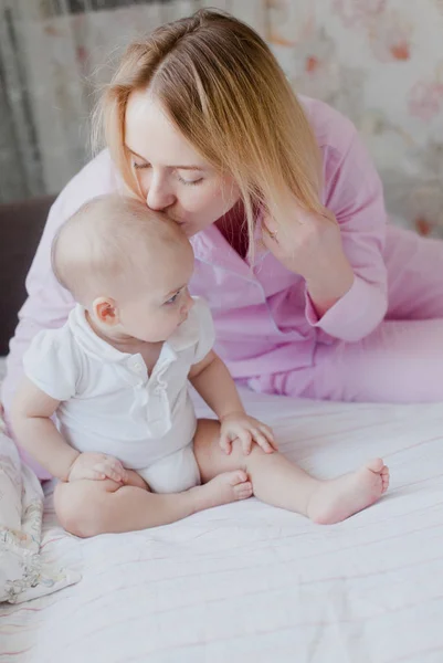 Mutter Blondine im rosa Pyjama küsst ihren Säugling auf ihrem Bett sitzend Stockfoto