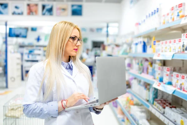 Sexy farmacéutico rubio en el campo médico utilizando la tecnología de computadora portátil y tableta para publicar anuncios de drogas en línea . — Foto de Stock
