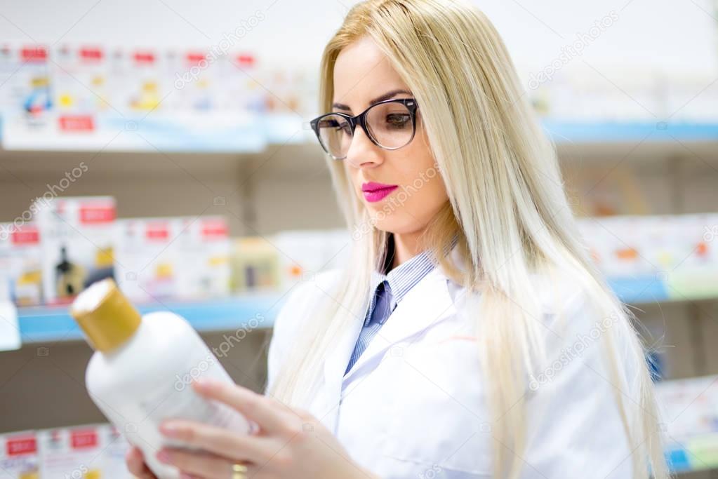 Young blonde pharmacist looking at pills and creams in store. Female medical assistant reading on labels