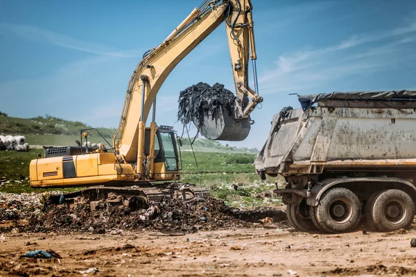 Industrial excavator using scoop and moving earth and trash at garbage dumping site.